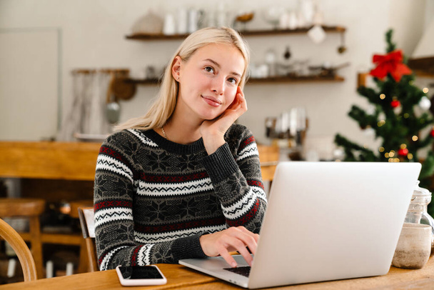 Image of a young dreaming smiling woman indoors at home using laptop computer with christmas tree on a background - Foto, Bild