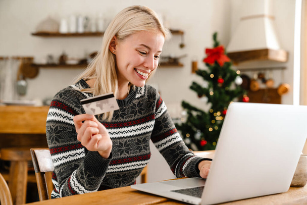 Photo of a young cheerful woman indoors at home using laptop computer and holding credit card - Фото, изображение