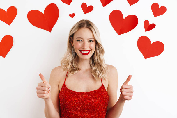 Photo of a young positive blonde woman in red dress showing thumbs up gesture isolated over white background with hearts - Fotó, kép