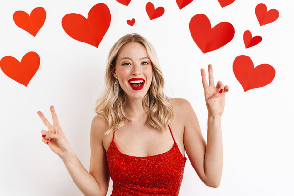 Photo of a young positive blonde woman in red dress showing peace gesture isolated over white background with hearts - Foto, Imagem
