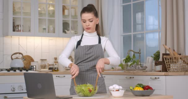 Mujer joven en delantal de cocina ensalada en la cocina, viendo video receta en el ordenador portátil en línea - Imágenes, Vídeo