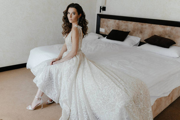 Mittleres Porträt einer fröhlichen, jungen Braut im Brautkleid, die auf dem Bett sitzt. Glückliches Mädchen posiert drinnen. - Foto, Bild