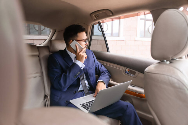 Homme d'affaires prospère avec ordinateur portable parlant par téléphone mobile dans une voiture moderne - Photo, image