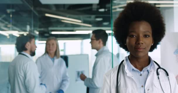 Close up portrait of young African American female professional doctor standing in hospital and smiling at camera. Caucasian doctors speaking on background. Healthcare concept - Footage, Video