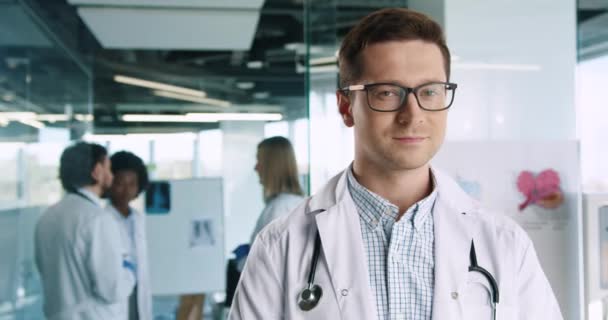 Close up portrait of Caucasian handsome happy young male professional doctor in glasses standing in hospital and smiling at camera. Doctors speaking on background. Healthcare concept - Footage, Video