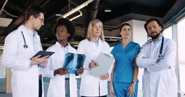 Grupa mieszanych ras zawodowych lekarzy pielęgniarki i specjaliści patrząc na aparat fotograficzny w dobrym nastroju, stojąc w nowoczesnym szpitalu ze skanowaniem rentgenowskim i tabletkami w rękach Koncepcja opieki zdrowotnej - Materiał filmowy, wideo
