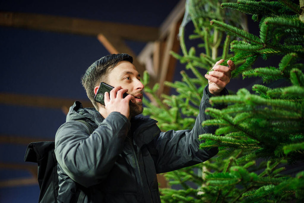 Festive shopping, selection and purchase of New Years attributes. Caucasian man speaks on phone and advises on choosing Christmas tree on evening of New Year Eve. Male buys spruce and uses smartphone. - Photo, Image