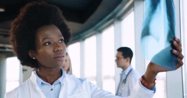 Ritratto ravvicinato di afroamericana giovane bella specialista sanitaria professionale femminile in piedi in ospedale moderno e guardando la scansione a raggi X. Medici che parlano sullo sfondo - Filmati, video