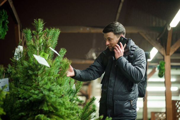 Young caucasian man choosing christmas tree in an outdoor market in evening talking on phone with his wife and consulting on right choice. Symbol of Christmas and New Year holidays. Holiday shopping. - Photo, Image