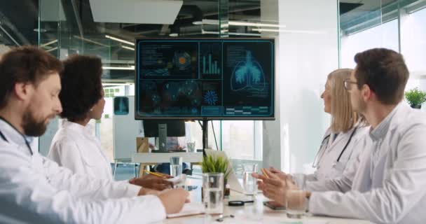 Medyczny wieloetniczny zespół lekarzy omawiający dane koronawirusa covid-19 na ekranie wyświetlacza cyfrowego pokazującego epidemię wirusa oraz infekcję ludzkiego ciała i płuc. Pojęcie medyczne - Materiał filmowy, wideo