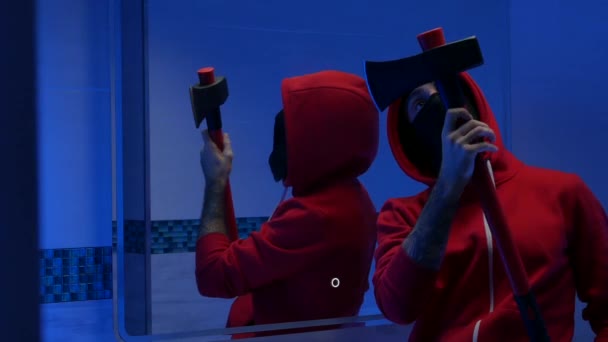een man met zijn gezicht bedekt in het toilet voor een spiegel met een brandbijl bereidt zich voor op een misdaad - Video