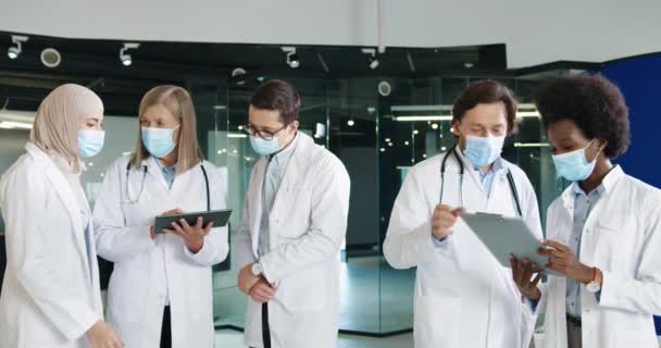 Wielu etnicznych lekarzy płci męskiej i żeńskiej pracownicy służby zdrowia w maskach medycznych dyskutują o zakażeniu koronawirusem, pisząc na tabliczce stojącej w klinice. Koncepcja portretu. Pandemia wywołana przez wirus Covid-19 - Materiał filmowy, wideo