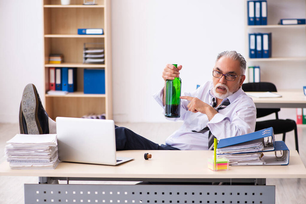 Old male employee drinking alcohol in the office - Фото, изображение