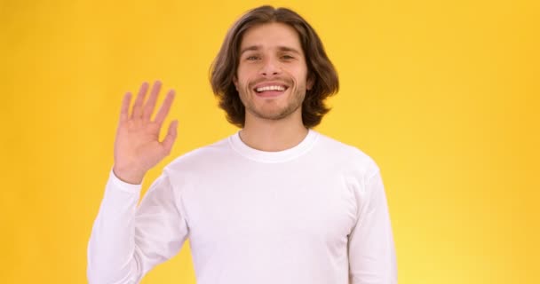 Студийный кадр молодого дружелюбного болтливого парня, машущего рукой и говорящего: Привет, замедленная съемка, оранжевый фон - Кадры, видео