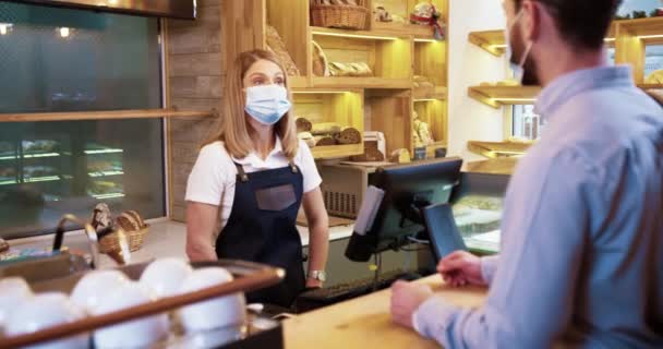 Καυκάσια υπάλληλος αρτοποιείου με ιατρική μάσκα στέκεται στη δουλειά και μιλάει με αρσενικό πελάτη που πουλάει ψωμί. Η ιδέα των μικρών επιχειρήσεων. Γυναίκα με ποδιά στον πάγκο στο αρτοποιείο. Έννοια τροφίμων - Πλάνα, βίντεο
