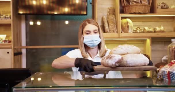 Sağlık maskeli güzel beyaz kadın satıcının portresi fırında çalışıyor fırın tezgahında taze ekmek tepsisiyle tepsiyi yerleştiriyor, kameraya iyi haldeki fırın konseptiyle bakıyor. - Video, Çekim