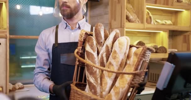 Крупным планом кавказского счастливого улыбающегося молодого продавца пекаря в защитном щите и перчатках, держащего корзину со свежими хлебными багетами, стоящими в пекарне в карантине. Концепция малого бизнеса - Кадры, видео