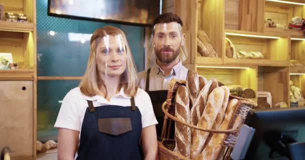 Portrait of Caucasian happy young married couple bakers in protective face shields and gloves holding basket with fresh bread standing in small own bakehouse in quarantine. Family business concept - Footage, Video