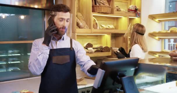 Jeune homme beau vendeur dans le bouclier facial debout au comptoir dans la boulangerie et parlant sur smartphone en prenant l'ordre du client par téléphone portable tandis que sa collègue féminine tapant sur la tablette derrière lui - Séquence, vidéo