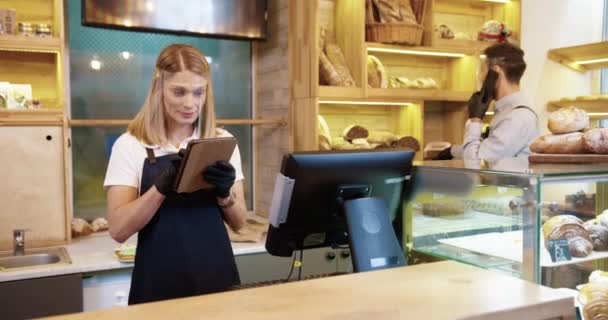 Fiatal gyönyörű kaukázusi nő eladó arc pajzs áll a pultnál pékségben és gépelés táblagépen, míg a kollégája férfi munkavállaló beszél a mobiltelefon rendelés ügyfél mögött - Felvétel, videó