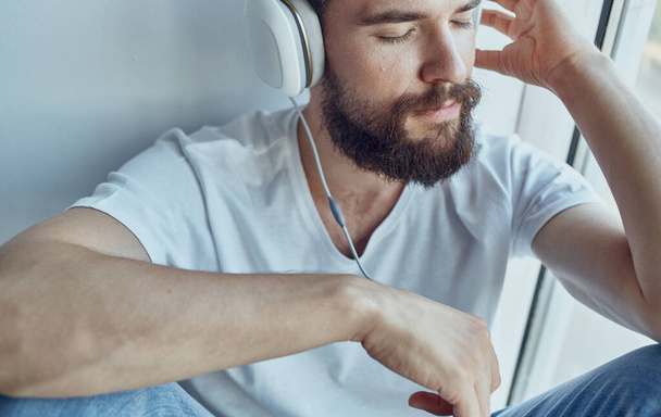 бородатый мужчина в наушниках слушает музыку возле окна весело - Фото, изображение