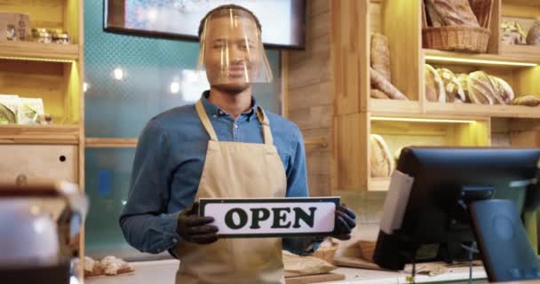 Portrait of cheerful African American young man seller in protective face shield and gloves holding Closed card in hands standing in bakehouse. Male baker reopening bakery shop. Small business concept - Footage, Video