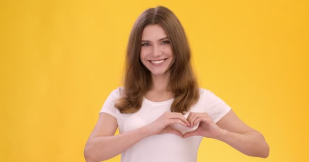 jonge gelukkige vrouw tonen hart gebaar, bewegen op haar borst en kijken door het, oranje studio achtergrond - Video