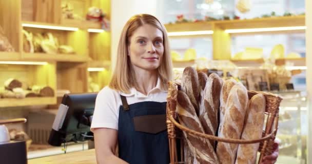 Закріпіть портрет щасливої молодої кавказької вродливої жінки, яка продає фартух у кошику зі свіжим хлібом у руках, стоячи у маленькому хлібному будинку, дивлячись на фотоапарат і посміхаючись поодинці. Концепція роботи - Кадри, відео