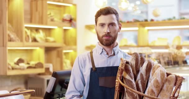 Ritratto ravvicinato di felice giovane caucasico bell'uomo barbuto venditore in grembiule cesto con pane appena sfornato in piedi in panetteria, guardando la fotocamera e sorridendo da solo. Concetto di lavoratore - Filmati, video