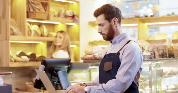 Close up of handsome young Caucasian man seller in apron standing in bakery, tapping and typing on laptop talking to colleague who works on background. Bakehouse concept. Small business - Footage, Video