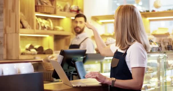 Close up of beautiful young Caucasian woman seller in apron standing in bakery, tapping and typing on laptop talking to male colleague working on background. Bakehouse concept. Small business - Footage, Video