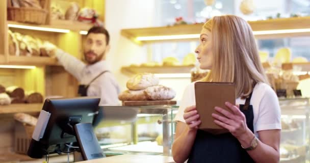Lähikuva nuori valkoihoinen kaunis naispuolinen myyjä esiliina seisoo leipomossa, napauttamalla ja kirjoittamalla tabletin laitteen, kun taas miespuolinen kollega työskentelee taustalla. Bakehouse-käsite - Materiaali, video