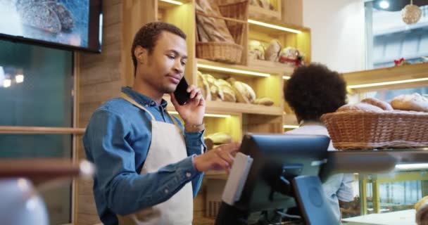 Крупным планом счастливого молодого привлекательного чернокожего рабочего-мужчины в фартуке разговаривающего по мобильному телефону, стоящего в пекарне, в то время как коллега-женщина работает, кладя хлеб на полки. Концепция предпринимательства - Кадры, видео