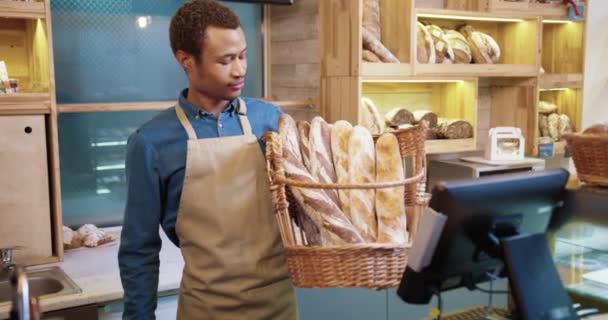 幸せな笑みを浮かべてハンサムなアフリカ系アメリカ人の若者起業家エプロン自分の小さなパン屋の手に新鮮なパンのバゲットを保持し、カメラを見て立っている。肖像画の概念 - 映像、動画