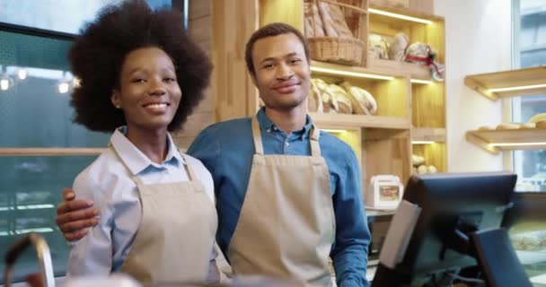 Portrait of joyful African American couple running family business. Man and woman bakers in aprons stands in bakehouse and smiling to camera hugging together. Small own bakery shop. Business concept - Footage, Video