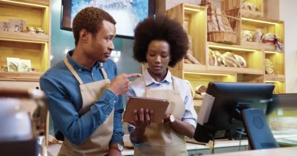 Portrait of happy smiling young African American couple man and woman in aprons using tablet tapping and typing while standing in own bakery shop talking indoor. Bakehouse concept - Footage, Video