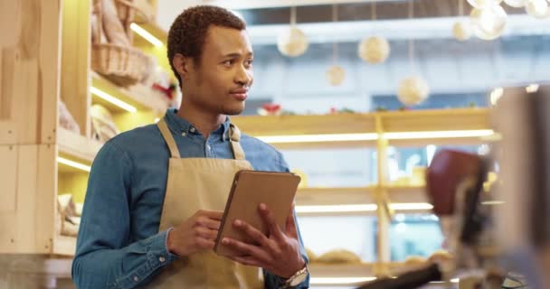 Крупным планом афроамериканец счастливый молодой мужчина, работающий в фартуке, стоит в булочной, пишет смс и печатает на планшете. Мужской продавец просматривает онлайн на устройстве. Бизнес-концепция. Розничная промышленность - Кадры, видео
