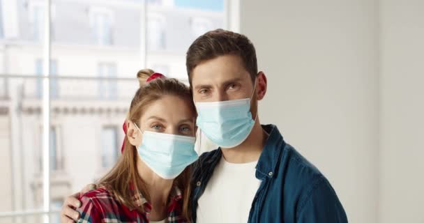 Közelkép portré boldog kaukázusi fiatal házaspár férfi és nő orvosi maszk áll az új lakás ölelés és nézi a kamerát. Felújítási koncepció. Újratervező szoba. Háztartási javítás - Felvétel, videó