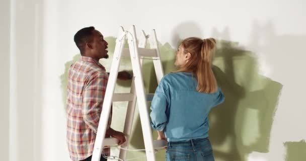 Hinter gemischten Rassen stehen ein afroamerikanischer Mann und eine kaukasische Frau auf einer Leiter und sprechen, während sie die Wände des Zimmers mit Pinselwalze in neuer Farbe reparieren und neu dekorieren. Erneuertes Konzept - Filmmaterial, Video