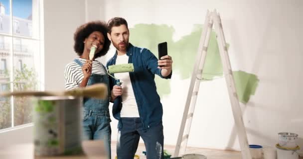 Neşeli karışık ırklar genç bir çift karı koca fırçalarla gülümseyip odada akıllı telefondan selfie çekiyorlar. Ev onarımı konsepti. Kadın ve erkek yenileme sırasında fotoğraf çekiyorlar. - Video, Çekim