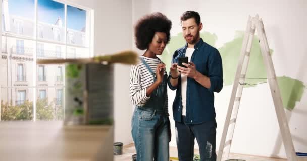 Joyful young married couple Caucasian husband and African American wife standing in room typing on smartphone choosing design of new home. Excited man and woman choosing decor for home renovation - Footage, Video