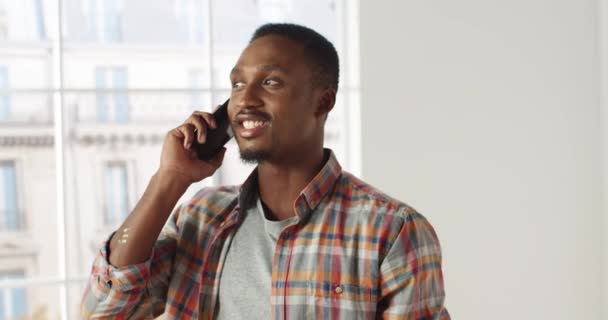 Κοντινό πλάνο του χαρούμενου Αφροαμερικανού όμορφου άντρα να χαμογελάει και να μιλάει στο κινητό σε διαμέρισμα με καλή διάθεση. Happy άνθρωπος καλώντας στο smartphone για την επισκευή στο σπίτι. Σχέδιο ανακαίνισης - Πλάνα, βίντεο