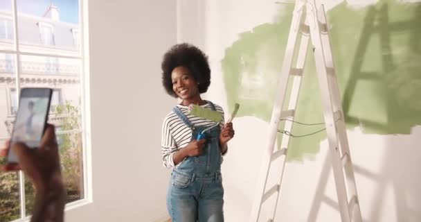 African American lovely husband taking photos of beautiful happy wife in room doing renovation in house posing with paint brush in hands. Renovation and home repair concept. Photoshoot on cellphone - Footage, Video