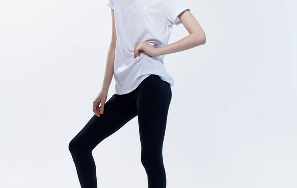 An athletic woman in a white T-shirt and leggings on a light background spread her legs to the sides - Foto, afbeelding