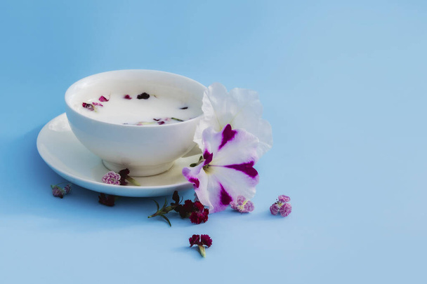 Fehér kupa vegán tejjel és virágokkal, kék háttérrel. Fogalma vegetáriánus italok és élelmiszerek, gyógynövény teák, szépség és egészség, Spa. - Fotó, kép