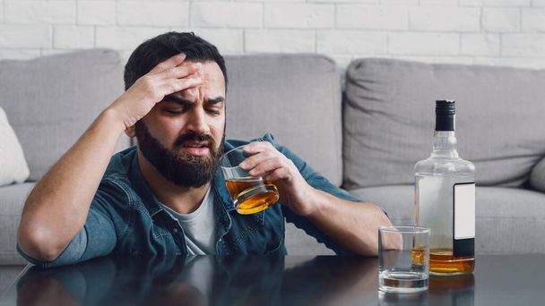 Πρόβλημα εξάρτησης από το αλκοόλ. Ένας θλιμμένος άντρας με γενειάδα κοιτάζει ένα ποτήρι ουίσκι. - Φωτογραφία, εικόνα