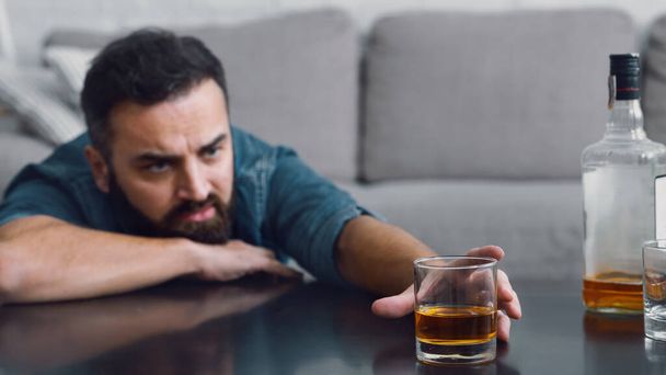 Frustrierter Mann mit Bart nimmt im Wohnzimmer ein Glas Whiskey vom Tisch - Foto, Bild