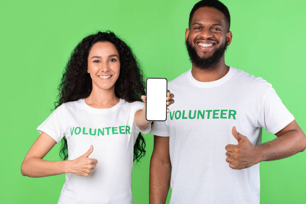 Два волонтера показывают телефон пустым экраном Жест, как, зеленый фон - Фото, изображение