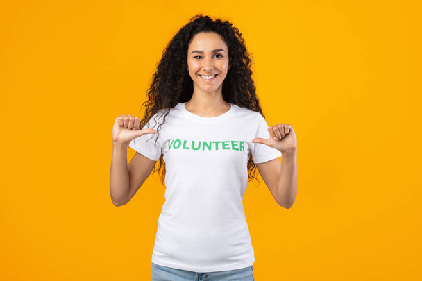 gelukkig vrouw vrijwilliger wijzend op zichzelf staande over gele achtergrond - Foto, afbeelding