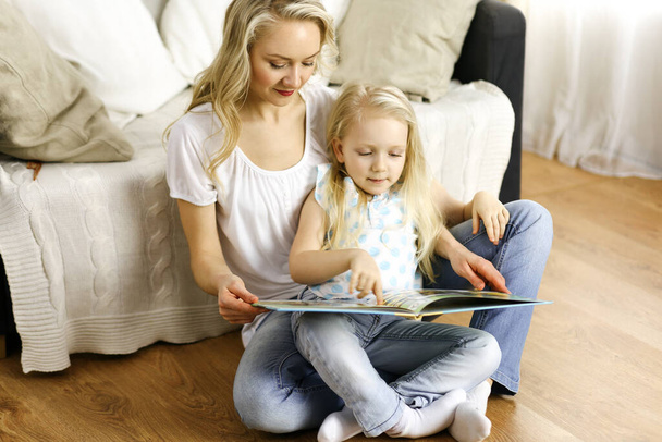 Happy family. Blonde young mother reading a book to her cute daughter while sitting at wooden floor. Motherhood concept - Foto, imagen